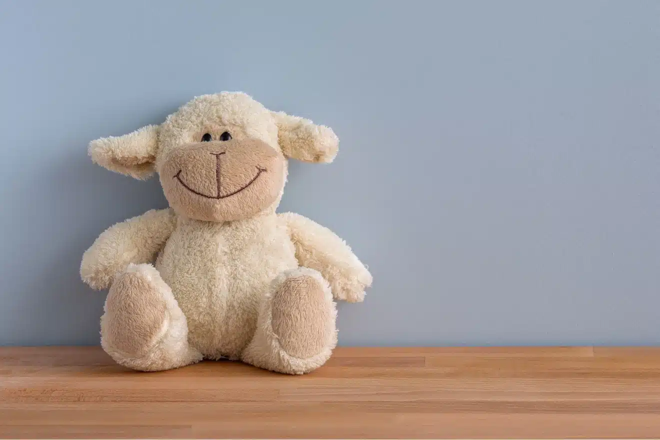 A plush sheep toy with a smiling face sits on a wooden surface against a plain light blue wall.