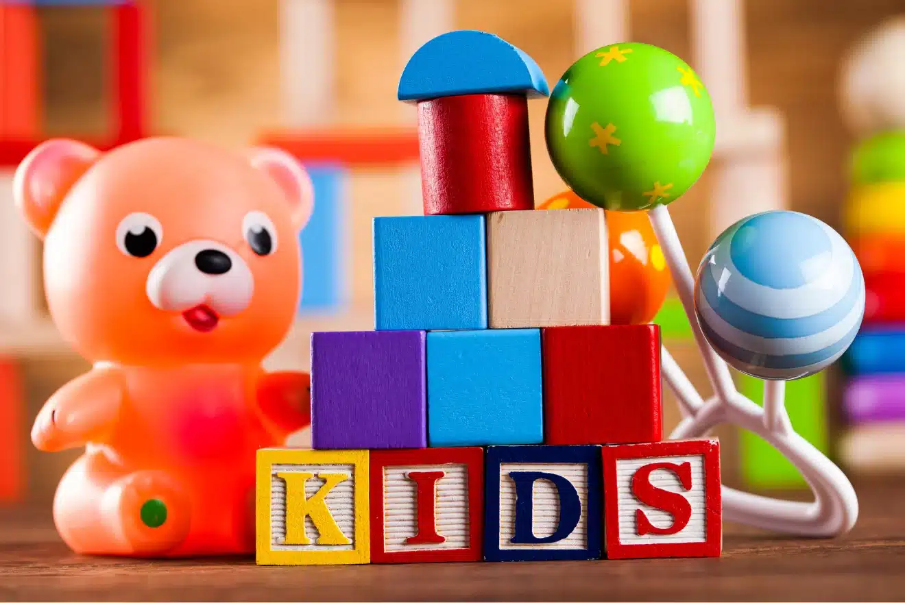 A plastic bear toy, colorful wooden blocks stacked with "KIDS" spelled out, and two baby rattles on a wooden surface.