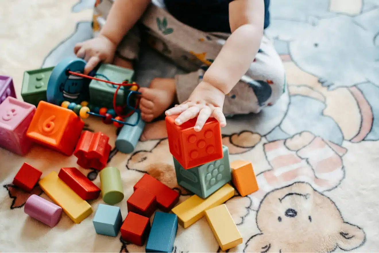 A child sits on a patterned rug, stacking colorful toy blocks and playing with various wooden and plastic toys.