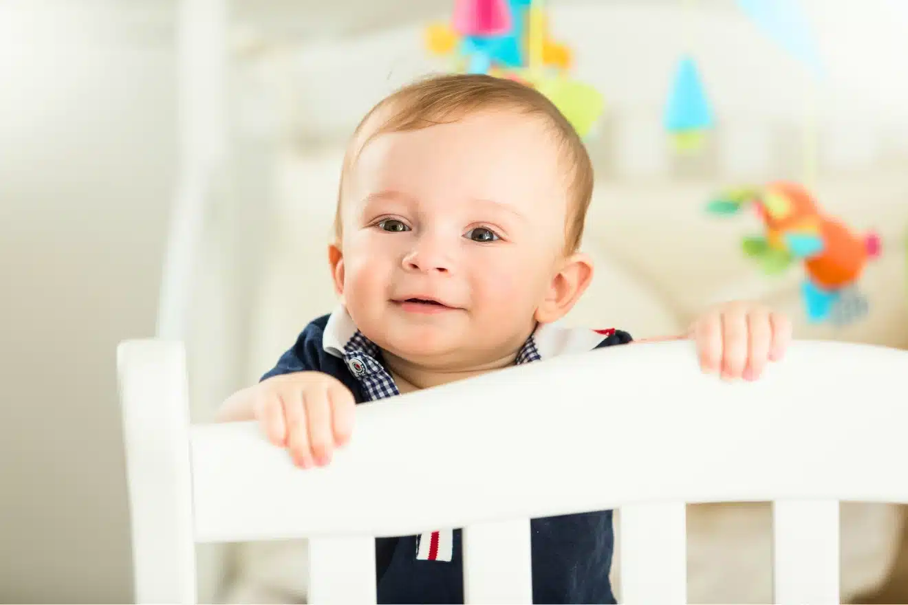 A baby stands in a white crib, holding onto the rail and looking outward, with colorful toys hanging in the background.