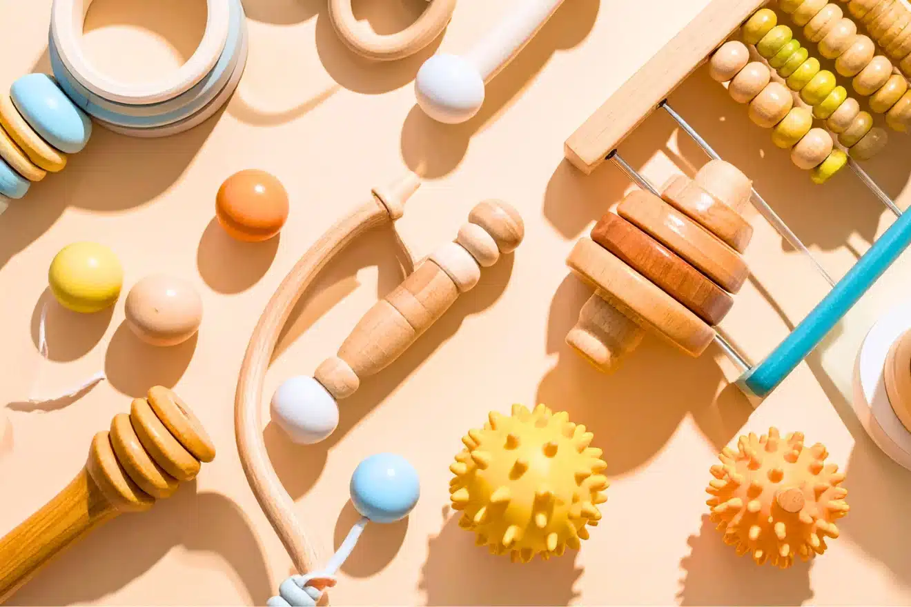 Assorted wooden and rubber baby toys, including rattles, rings, abacus, and textured balls, arranged on a light beige surface.