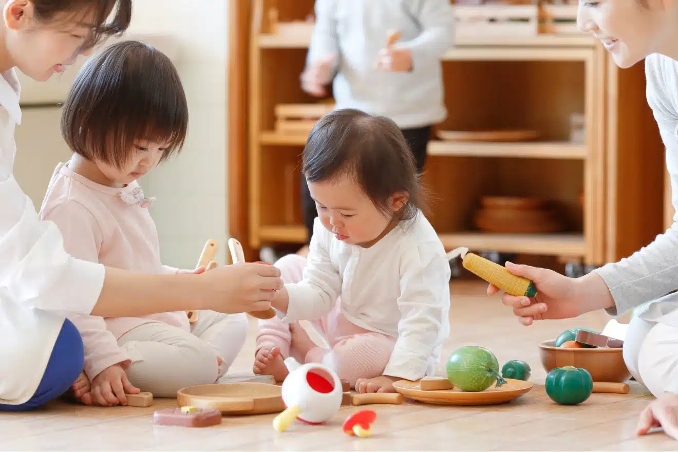 “Infants playing with wooden food toys in a Montessori classroom with caregivers — montessori toys for infants encouraging sensory exploration and cooperative play.