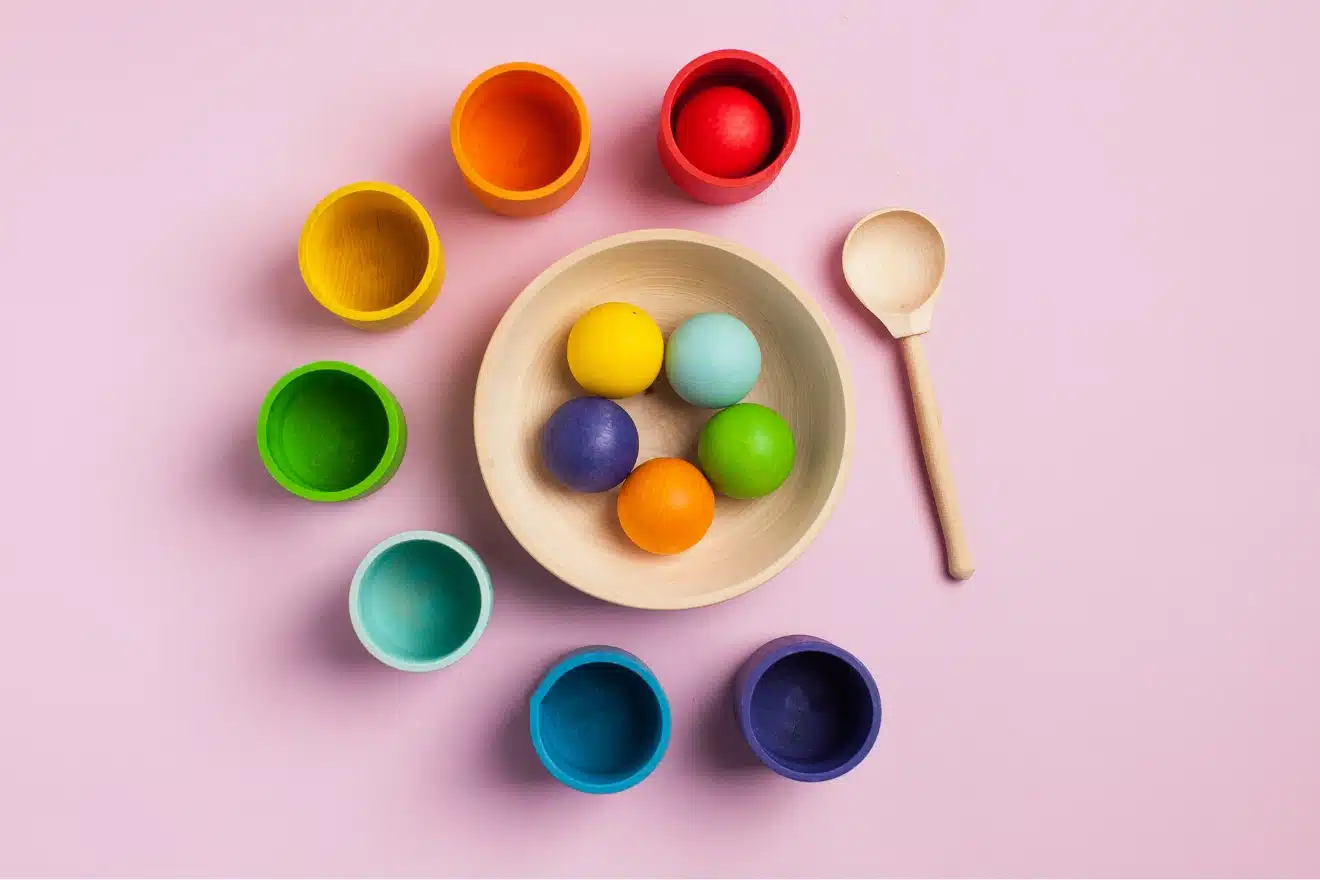 Colorful wooden cups and balls with a spoon on a pink background — montessori toys for infants that encourage color matching and sensory play.