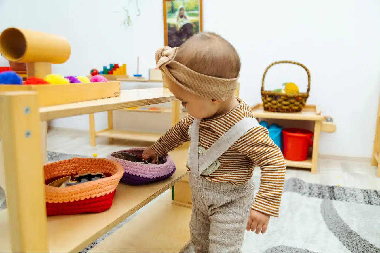 Infant exploring baskets of wooden and sensory toys on low shelves in a Montessori classroom — montessori toys for infants encouraging independent play and fine motor development.