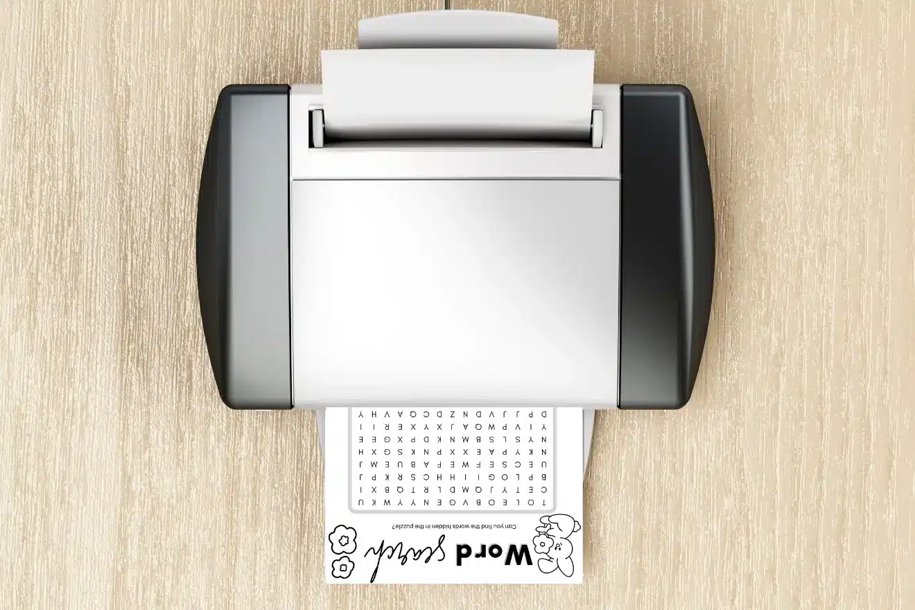 Top-down view of a printer on a wooden surface printing a word search puzzle with small flower illustrations at the bottom.